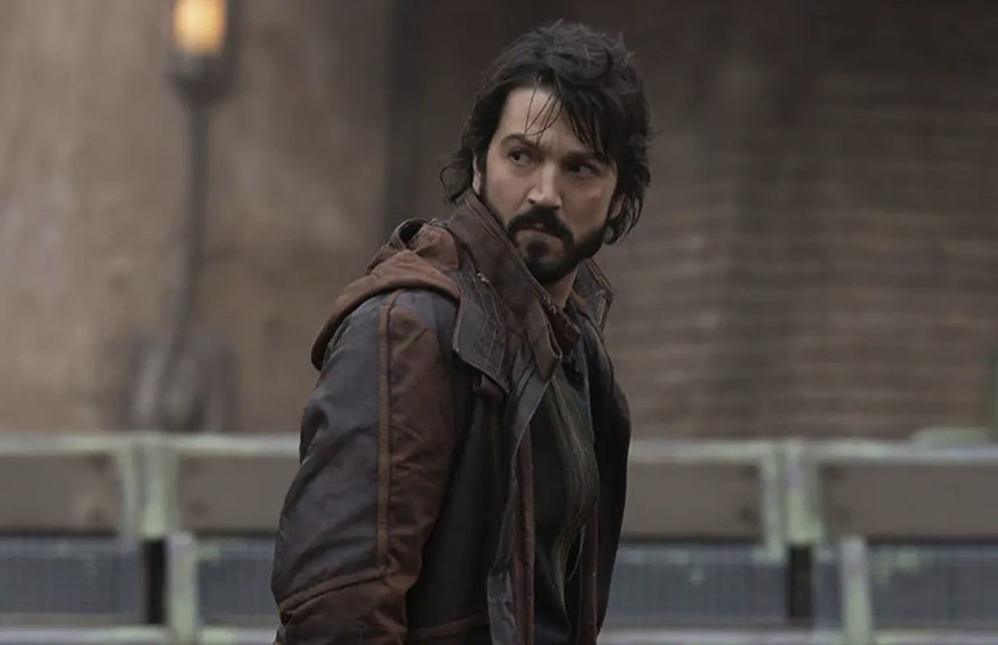 Cassian Andor is standing outside, in front of a brick building, looking over his shoulder.