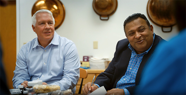 An elderly gentleman with white hair and a gentleman with darker skin and brown hair sit next to each other at a table talking with someone not pictured.