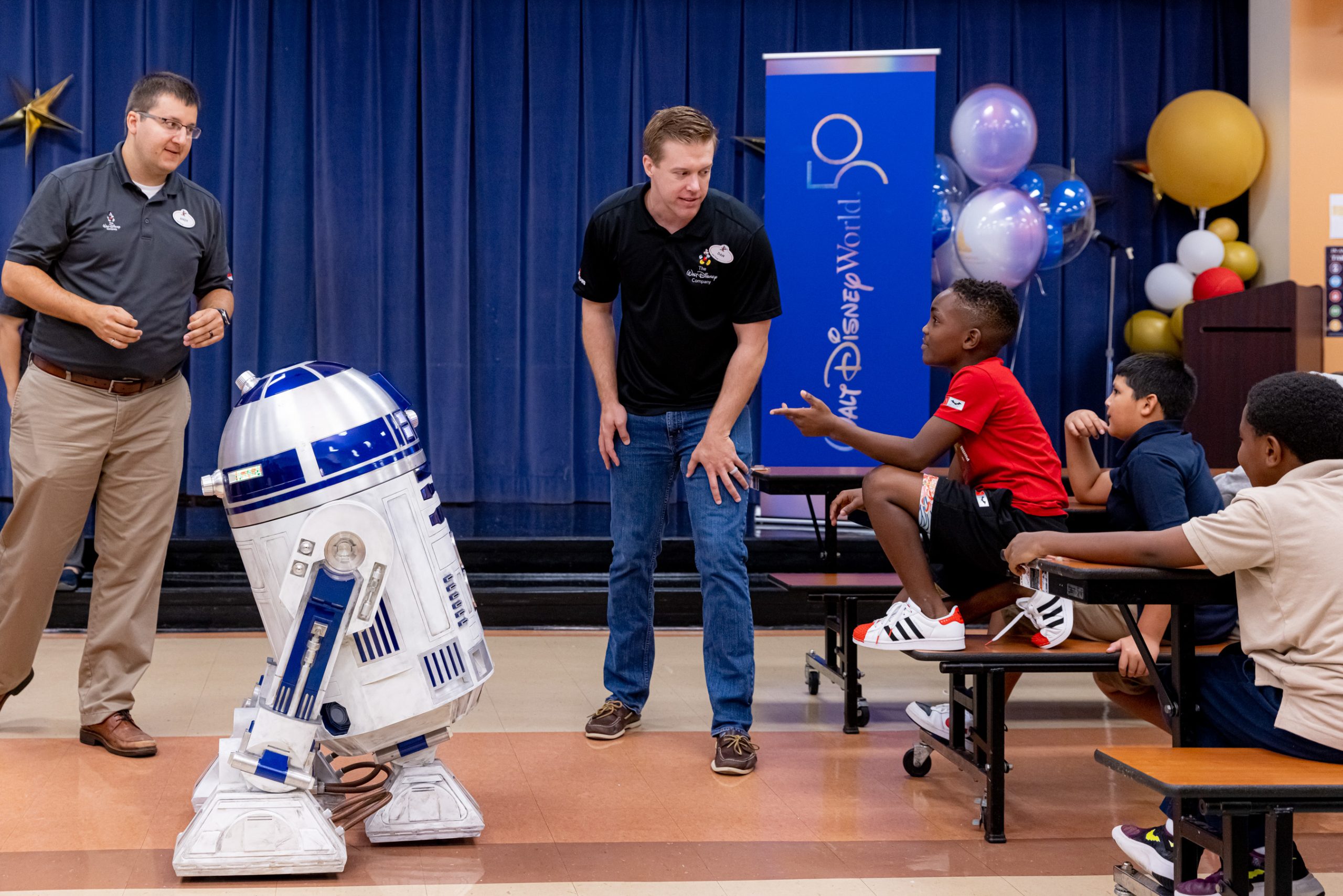 R2-D2 is in front of a Black child kneeling on a bench. The child is wearing a red shirt, black shorts and while athletic shoes pointing towards R2-D2.